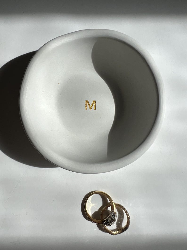 May include: A white, round, shallow dish with a gold glitter "M" in the center. Two gold rings with gemstones are next to the dish. The dish is likely a jewelry holder.