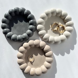 Op de afbeelding: Drie ronde betonnen schalen voor snuisterijen met een verhoogde, getextureerde rand. De schalen zijn grijs, wit en beige. De witte schaal heeft twee gouden oorringen erin. De beige schaal heeft een gouden ring erin.