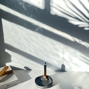 Op de afbeelding: Een smeulende Palo Santo-stok in een donkergrijze keramische wierookhouder. De houder is rond met een kleine rechthoekige rust. Een witte doos met de tekst "ABHAME IMAGE" staat links. Rook stijgt op tegen een witte achtergrond.