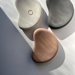 K&ouml;nnte beinhalten: Drei glatte, organisch geformte Beton-Tabletts in neutralen Farben: hellgrau, dunkelgrau und beige. Das hellgraue Tablett hat eine kleine kreisf&ouml;rmige Vertiefung. Die Tabletts sind auf einer wei&szlig;en Oberfl&auml;che mit Schatten angeordnet.