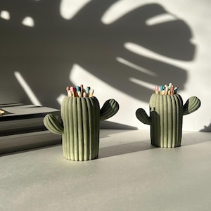 May include: Two green cactus shaped matchstick holders with colourful matchsticks inside. The holders are on a white surface.