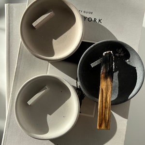 Op de afbeelding: Drie kleine, ronde betonnen asbakken in grijstinten en gebroken wit, elk met een rechthoekige rust. E&eacute;n asbak bevat een gedeeltelijk verbrande palo santo-stok. De asbakken liggen op een boek.