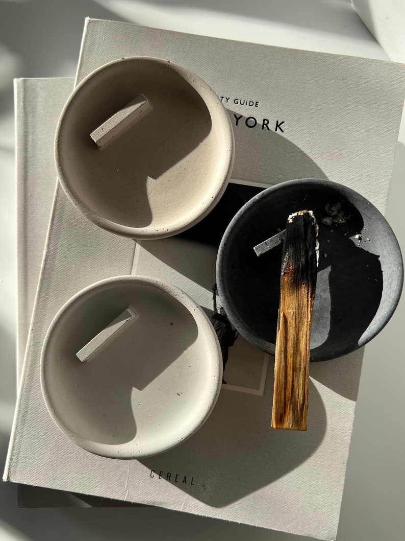 Op de afbeelding: Drie kleine, ronde betonnen asbakken in grijstinten en gebroken wit, elk met een rechthoekige rust. E&eacute;n asbak bevat een gedeeltelijk verbrande palo santo-stok. De asbakken liggen op een boek.
