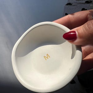 May include: A small, white, ceramic bowl with an irregular shape. The bowl has a smooth interior and a slightly textured exterior. A gold-colored emblem is visible at the bottom of the bowl. The bowl is held by a person with red nail polish.