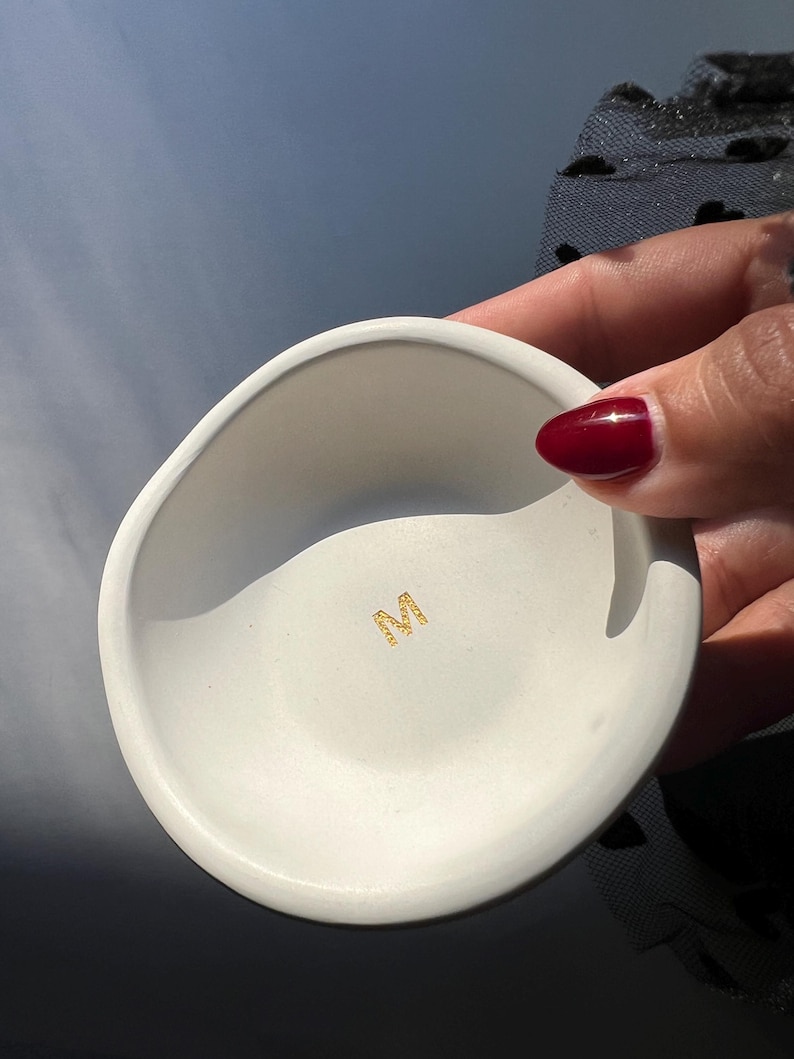 May include: A small, white, ceramic bowl with an irregular shape. The bowl has a smooth interior and a slightly textured exterior. A gold-colored emblem is visible at the bottom of the bowl. The bowl is held by a person with red nail polish.