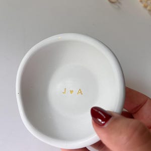 May include: A small, white ceramic ring dish with a slightly irregular shape. The dish features gold lettering spelling out "J ♡ A" in the center. The dish is held by a person with red nail polish.