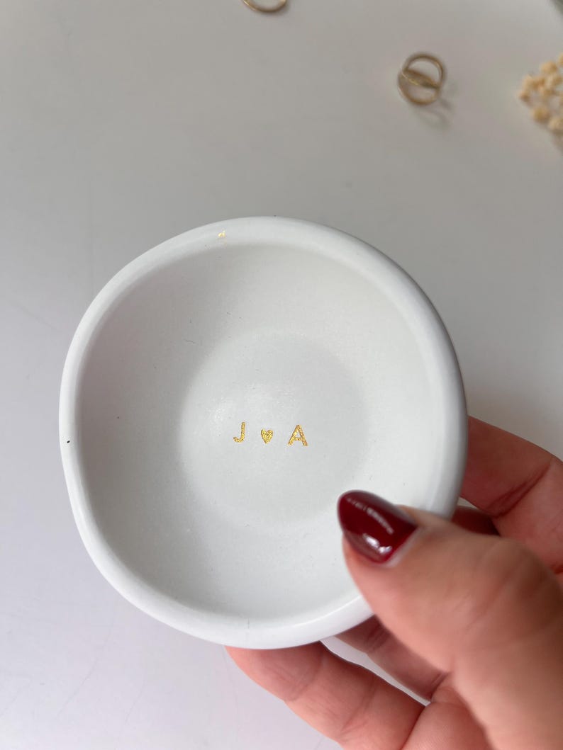 May include: A small, white ceramic ring dish with a slightly irregular shape. The dish features gold lettering spelling out "J ♡ A" in the center. The dish is held by a person with red nail polish.