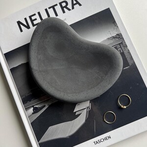 K&ouml;nnte beinhalten: Eine dunkelgraue, organisch geformte Schale liegt auf einem schwarz-wei&szlig;en Buch mit dem Titel "NEUTRA". Zwei goldene Ringe sind in der N&auml;he der Schale platziert. Das Buchcover zeigt ein Foto eines Geb&auml;udes.