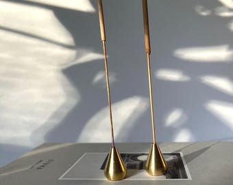 Könnte beinhalten: Zwei goldene Metall-Räucherstäbchenhalter in Kegelform. Jeder Halter hat einen braunen Räucherstäbchen in die Spitze gesteckt.