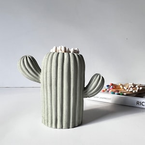 May include: A grey concrete cactus shaped matchstick holder with white matchsticks inside. The holder is sitting on a white surface with a book in the background.