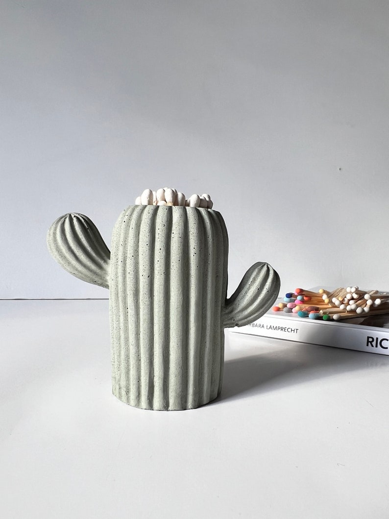 May include: A grey concrete cactus shaped matchstick holder with white matchsticks inside. The holder is sitting on a white surface with a book in the background.