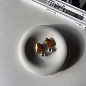 May include: A pair of gold and silver earrings with a hammered texture. The earrings are displayed on a white ceramic dish.