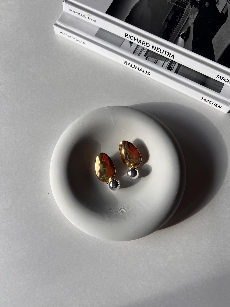 May include: A pair of gold and silver earrings with a hammered texture. The earrings are displayed on a white ceramic dish.