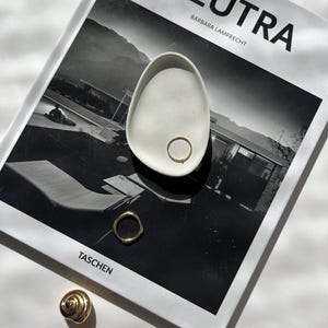 Peut inclure: Un plat à bagues en céramique blanche de forme arrondie, posé sur un livre de photographie en noir et blanc intitulé "NEUTRA BARBARA LAMPRECHT TASCHEN". Trois bagues en or sont exposées sur le plat et sur le livre.