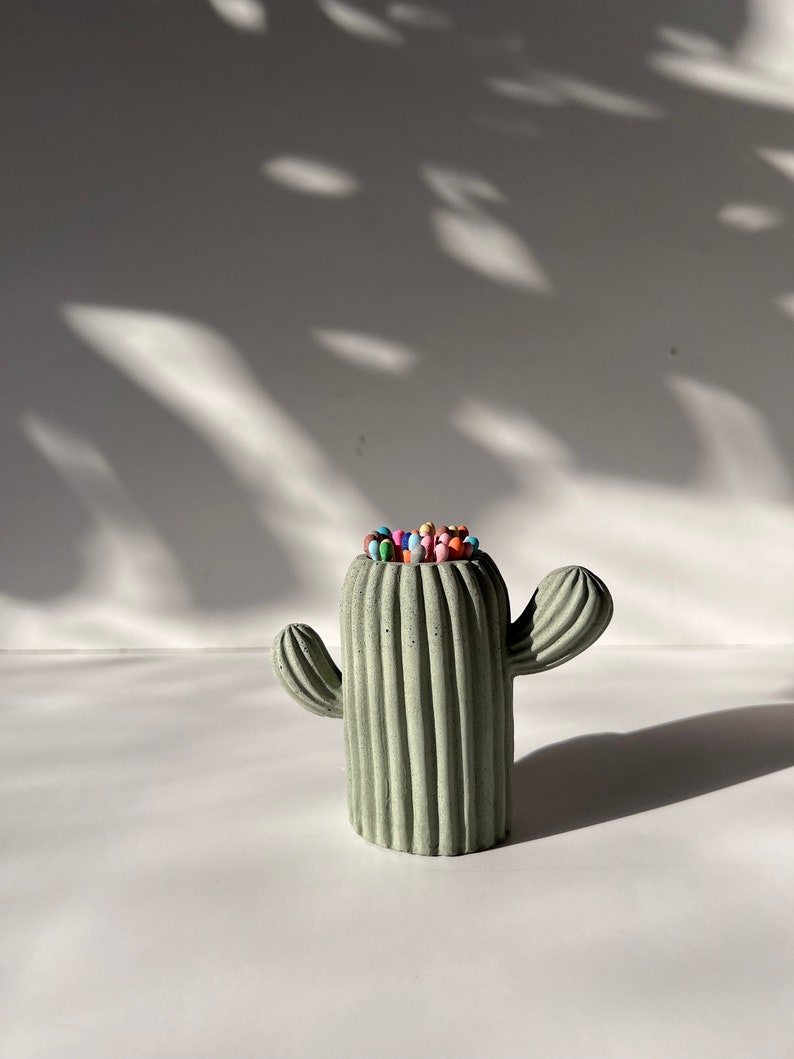 May include: A green concrete cactus shaped matchstick holder with colourful matchsticks inside.