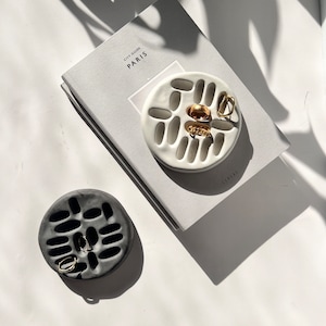 May include: Two ceramic ring dishes, one white and one black, with a variety of gold rings in the white dish. The dishes are on a white surface with a book titled "City Guide Paris" in the background.