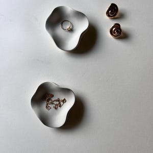 Peut inclure: Deux plats en c&eacute;ramique blanche aux bords ondul&eacute;s, l'un avec une bague en or et l'autre avec un collier en cha&icirc;ne dor&eacute;e. Les plats sont sur une surface blanche.