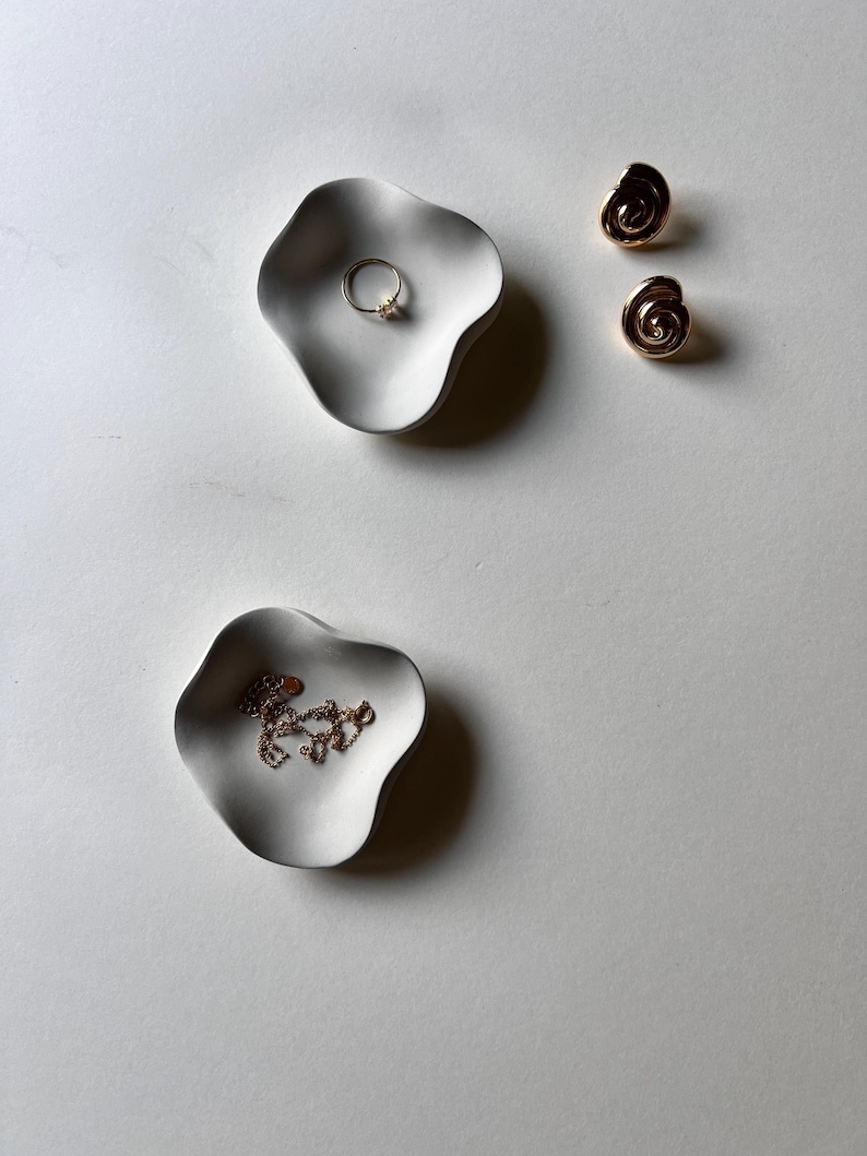 Peut inclure: Deux plats en c&eacute;ramique blanche aux bords ondul&eacute;s, l'un avec une bague en or et l'autre avec un collier en cha&icirc;ne dor&eacute;e. Les plats sont sur une surface blanche.
