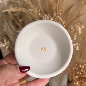 May include: A small, white ceramic dish with a gold letter "M" in the center. The dish is round with a slightly raised edge. The background is blurred, with dried plants and a neutral color palette.