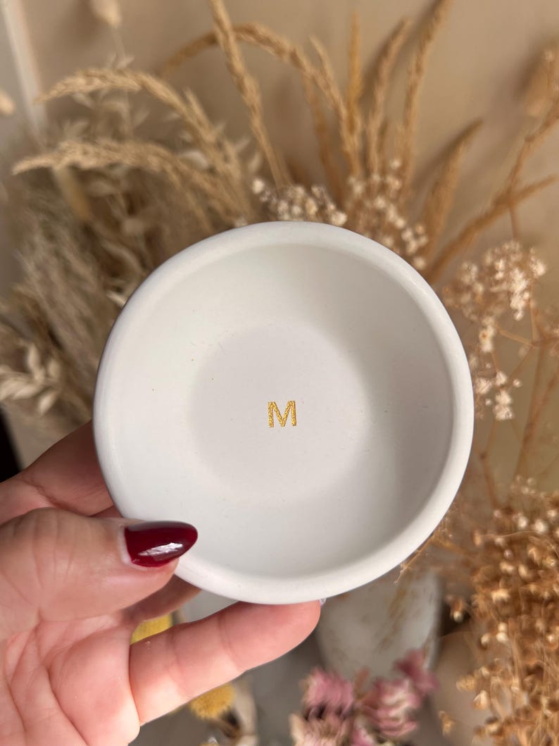 May include: A small, white ceramic dish with a gold letter "M" in the center. The dish is round with a slightly raised edge. The background is blurred, with dried plants and a neutral color palette.