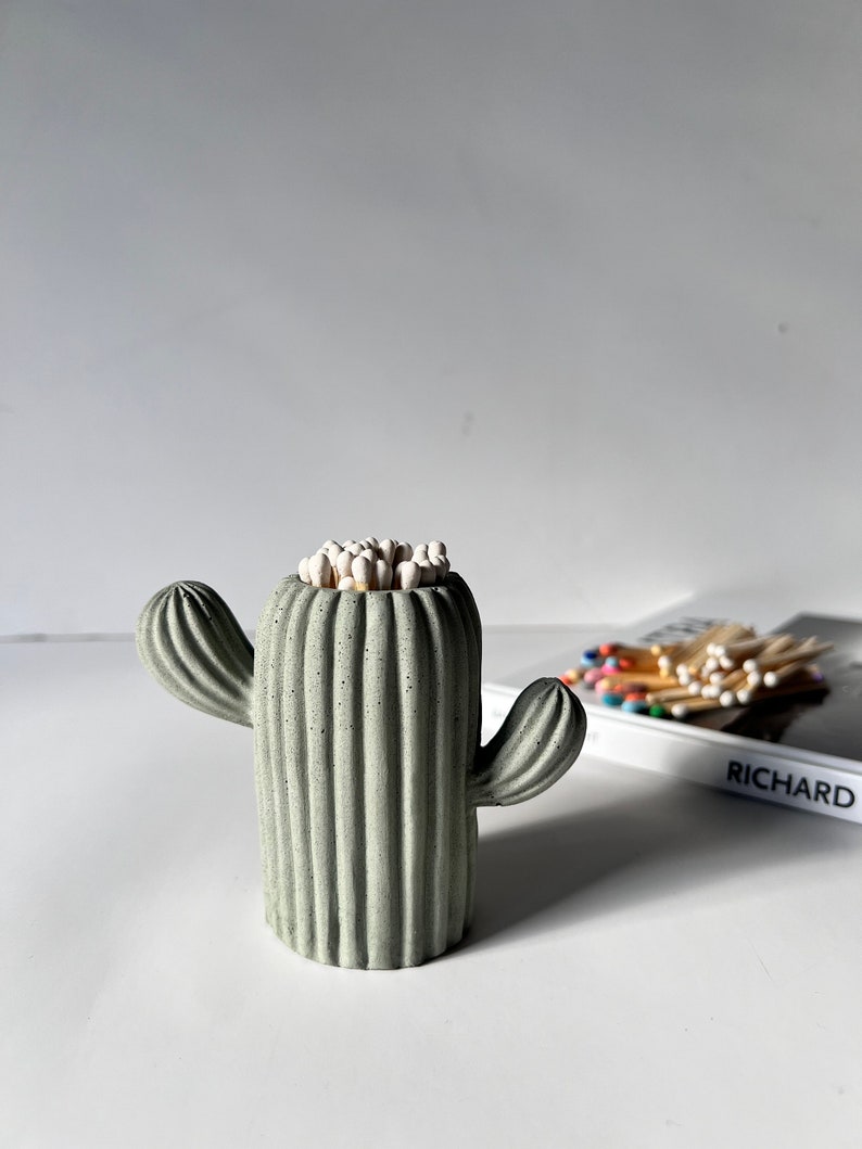 May include: A green ceramic cactus shaped matchstick holder with white matchsticks inside. The holder is sitting on a white surface.