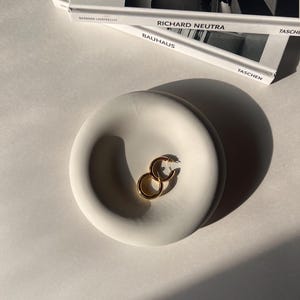 May include: A white ceramic dish with a rounded, indented center. Two gold hoop earrings are resting in the dish.
