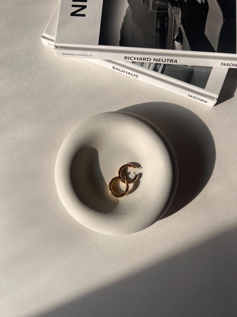 May include: A white ceramic dish with a rounded, indented center. Two gold hoop earrings are resting in the dish.