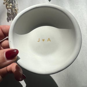 May include: A small, round, white ceramic dish with gold lettering of the initials "J & A" and a heart symbol. A gold ring is visible in the background. The dish is held by a hand with red nail polish.