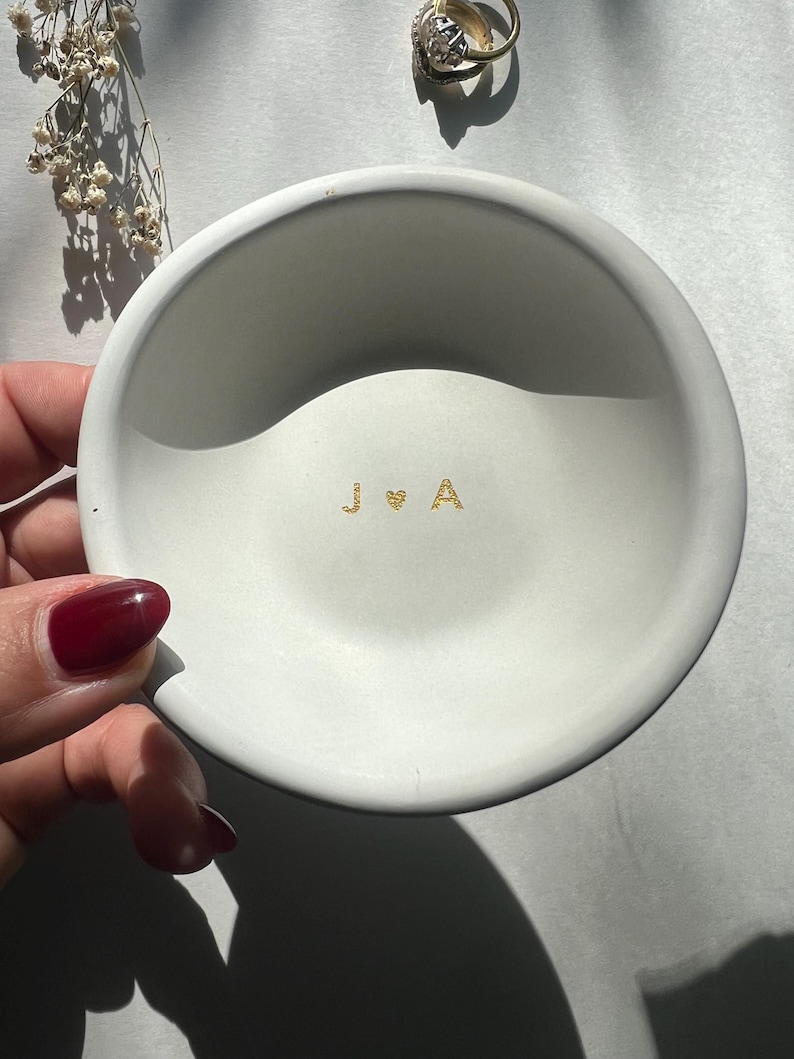 May include: A small, round, white ceramic dish with gold lettering of the initials "J & A" and a heart symbol. A gold ring is visible in the background. The dish is held by a hand with red nail polish.