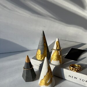May include: Four concrete ring holders in the shape of cones. The cones are painted with gold accents and are displayed on a white surface with the word "CEREAL" visible. One cone is on a book with the text "NEW YORK" visible.