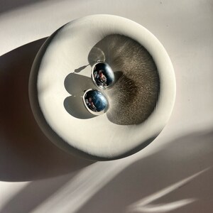 May include: Two silver oval-shaped earrings with a smooth, reflective surface. The earrings are displayed on a white ceramic dish.