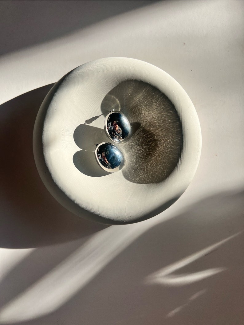 May include: Two silver oval-shaped earrings with a smooth, reflective surface. The earrings are displayed on a white ceramic dish.