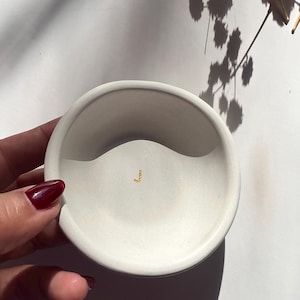 Op de afbeelding: Een kleine, crèmekleurige, ronde keramische schaal met een golvend ontwerp. In het midden bevindt zich een klein goudkleurig accent. De schaal wordt vastgehouden door een hand met donkerrode nagellak. De achtergrond is wit met schaduwen.