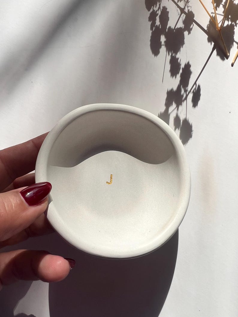 May include: A small, off-white, round ceramic dish with a wavy design. A small gold-colored accent is in the center. The dish is held by a hand with a dark red nail polish. The background is white with shadows.