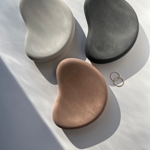 K&ouml;nnte beinhalten: Drei betonfarbene Tabletts in Form von Kieselsteinen in Grau, Schwarz und Beige. Zwei goldene Ringe liegen in der N&auml;he des beigefarbenen Tabletts. Die Tabletts haben eine glatte, strukturierte Oberfl&auml;che und sind auf wei&szlig;em Grund angeordnet.