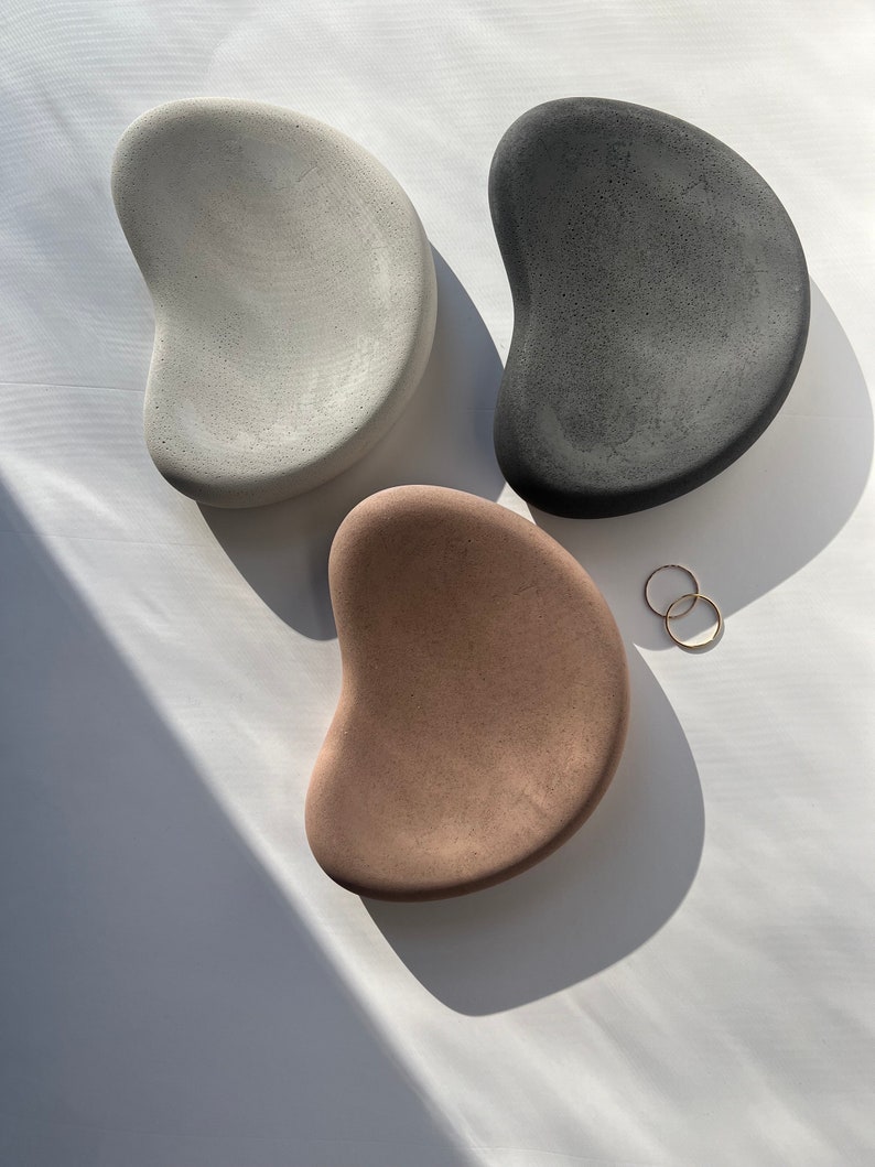 K&ouml;nnte beinhalten: Drei betonfarbene Tabletts in Form von Kieselsteinen in Grau, Schwarz und Beige. Zwei goldene Ringe liegen in der N&auml;he des beigefarbenen Tabletts. Die Tabletts haben eine glatte, strukturierte Oberfl&auml;che und sind auf wei&szlig;em Grund angeordnet.