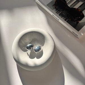 May include: Two silver, round, decorative earrings in a white ceramic dish. The dish has a rounded, organic shape.