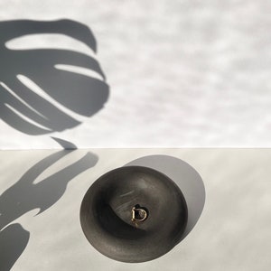 May include: A black ceramic bowl with a rounded shape and a gold ring inside. The bowl is sitting on a white surface.