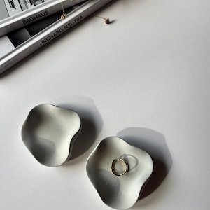 Peut inclure: Deux coupelles &agrave; anneaux en c&eacute;ramique blanche avec une forme ondul&eacute;e et organique. Une coupelle contient une bague en or.