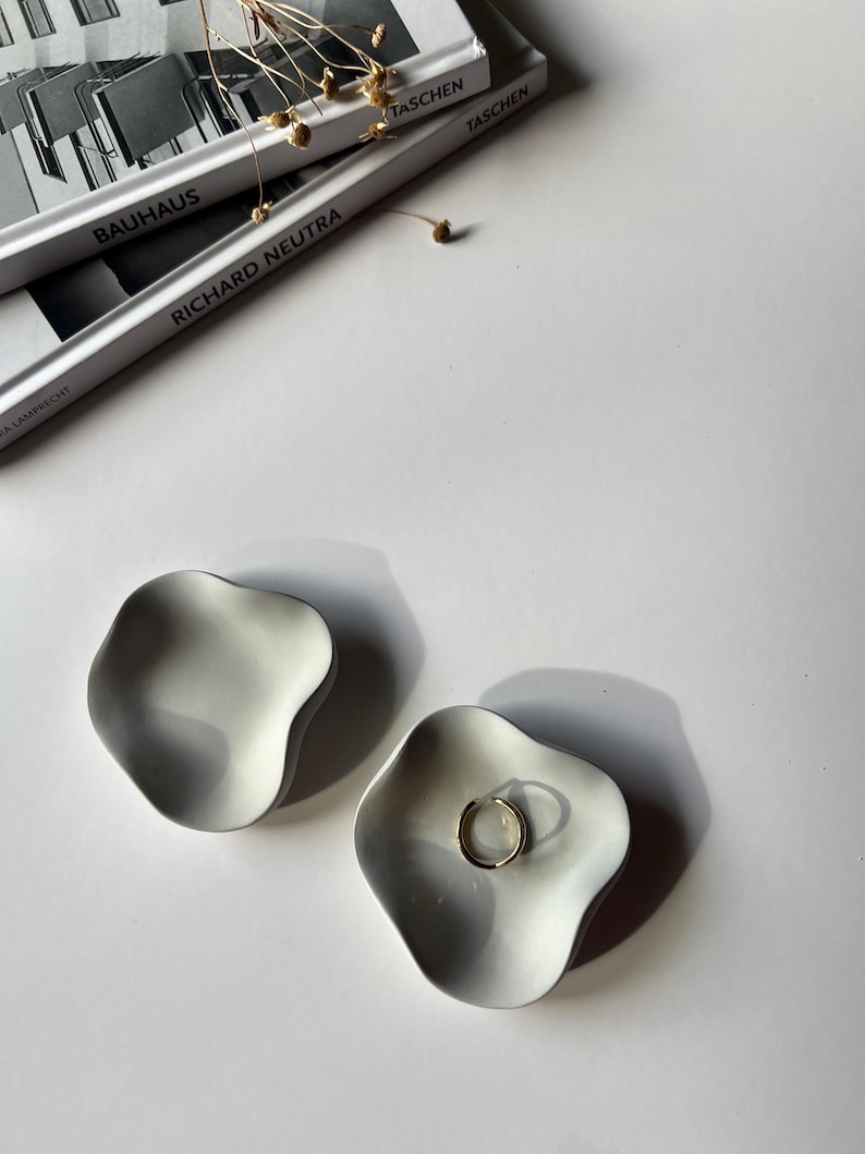Peut inclure: Deux coupelles &agrave; anneaux en c&eacute;ramique blanche avec une forme ondul&eacute;e et organique. Une coupelle contient une bague en or.