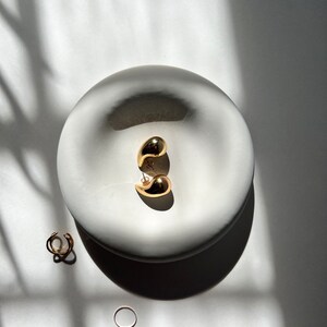 May include: A pair of gold teardrop shaped earrings in a white ceramic dish. There are two gold rings next to the dish.