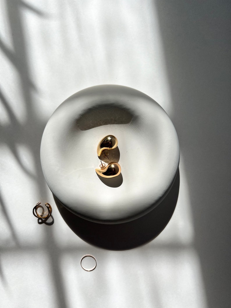 May include: A pair of gold teardrop shaped earrings in a white ceramic dish. There are two gold rings next to the dish.