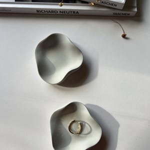 Peut inclure: Deux plats en c&eacute;ramique blanche avec une forme ondul&eacute;e et organique. Un plat contient une bague en or. Les plats sont sur une surface blanche.