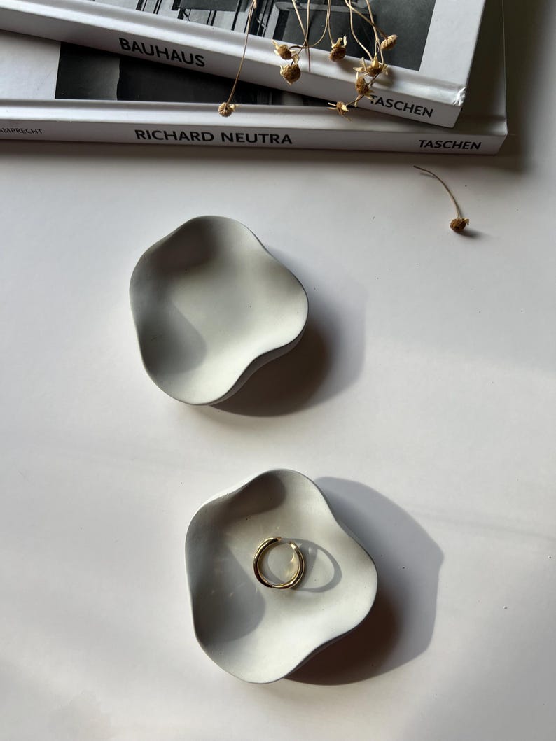Peut inclure: Deux plats en c&eacute;ramique blanche avec une forme ondul&eacute;e et organique. Un plat contient une bague en or. Les plats sont sur une surface blanche.