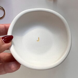 May include: A small, white, round ceramic dish with a shallow bowl. The dish has a gold-colored letter "J" in the center. Two gold rings are visible in the background. The dish is held by a hand with a red nail polish.