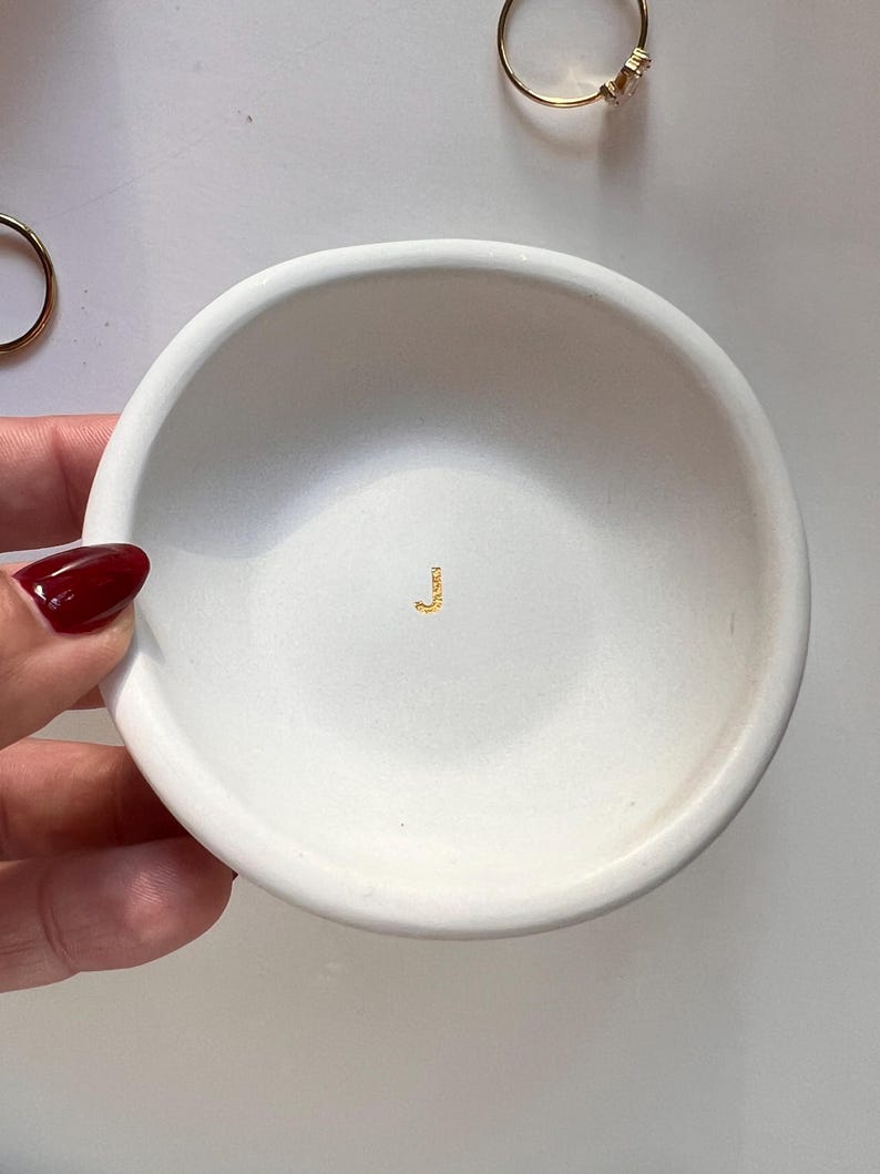 May include: A small, white, round ceramic dish with a shallow bowl. The dish has a gold-colored letter "J" in the center. Two gold rings are visible in the background. The dish is held by a hand with a red nail polish.