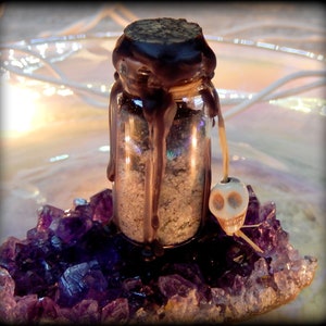 May include: A small glass bottle filled with a white substance and sealed with a dark brown wax. The bottle is sitting on a bed of purple amethyst crystals. A small white skull charm hangs from the bottle.