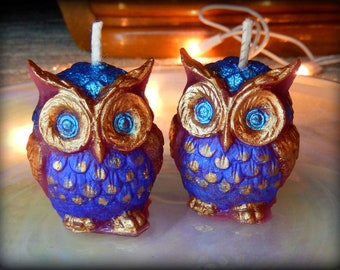May include: Two purple and gold owl candles with blue eyes and gold accents. The candles are on a white surface.