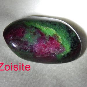 May include: A smooth, oval-shaped zoisite gemstone with a mix of green, pink, and black colors. The stone is polished and has a glossy finish. Zoisite.