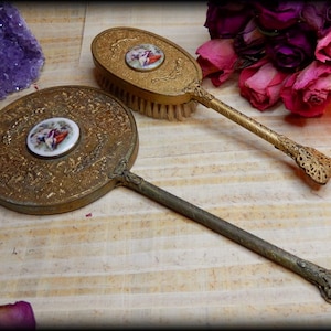 May include: Two antique gold-toned hand mirrors with ornate handles and a porcelain design in the center of each mirror. The mirrors are on a wooden surface.